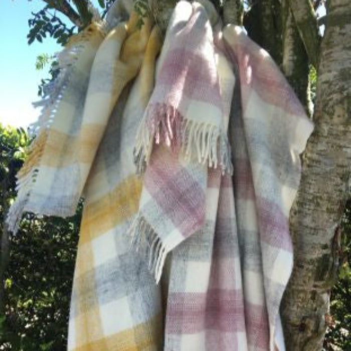 Two plaid wool blankets draped over a tree branch with a natural background, featuring colours of yellow, pink and grey