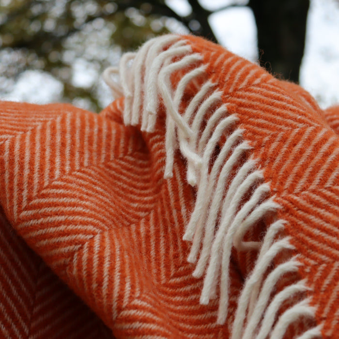 Pumpkin orange fishbone style wool blanket with white fringe against a blurred natural background