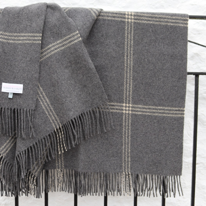 A dark gray luxury cashmere wool throw blanket with an off white windowpane check pattern and fringed edges, draped over a black metal railing against a white textured wall.