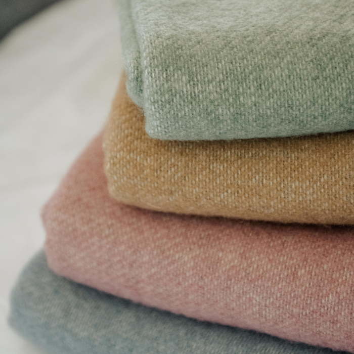 Stack of folded pure woollen blankets in muted shades of green, brown ochre, pink and grey blue on a light background. 