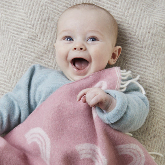 Baby lying on a textured surface with a Rainbow Pink Merino Wool Baby Blanket featuring white fringe