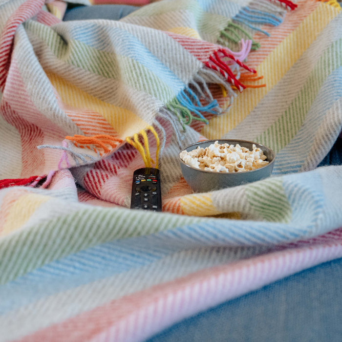 Colourful rainbow style herringbone wool blanket draped over a blue couch with a bowl of popcorn and remote control on top.