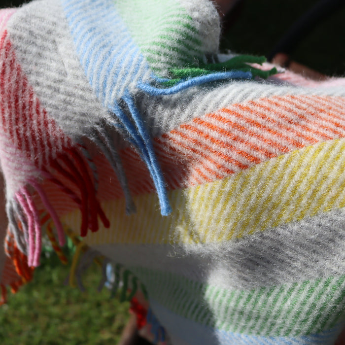 Close up of a colourful herringbone style , rainbow stripe pure wool blanket with fringe, with a blurred grassy background