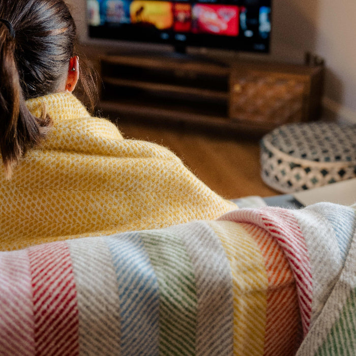 Person sitting with a colourful rainbow stripe blanket draped over the back of the couch, watching TV in a living room.