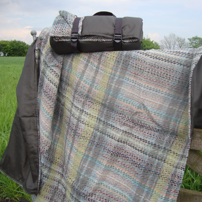 Plaid recycled blanket draped over a wooden fence with a rolled up blanket on top in a grassy field and trees in the background