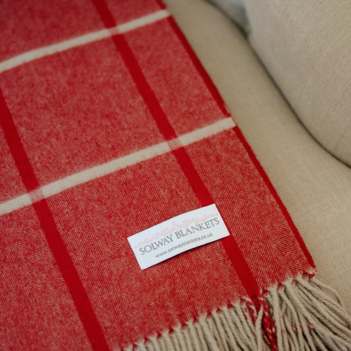 A close-up of a red and french grey plaid cashmere wool blanket folded on a beige upholstered armchair, with a white brand label reading 'SOLWAY BLANKETS' visible near the bottom.