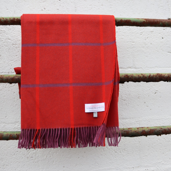 A red woven cashmere blanket throw with purple stripes and fringed edges, draped over a rustic metal railing against a white textured wall. A white brand label reading 'SOLWAY BLANKETS' is visible near the bottom.