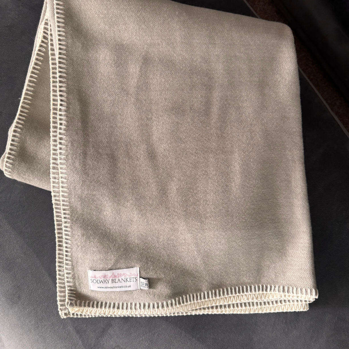 A folded dove grey cotton throw blanket with cream-colored blanket stitched edges and a visible 'Solway Blankets' brand label, laid flat on a dark grey surface.