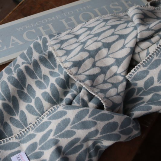 A close-up of a soft woven cotton throw blanket with a dusty duck egg blue and off-white leaf pattern, draped over a dark wooden surface next to a decorative sign.