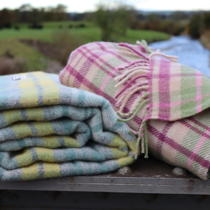 Two plaid woollen blankets, one with blue and green colourings and one with pink and green, on a wooden surface with a natural landscape in the background.