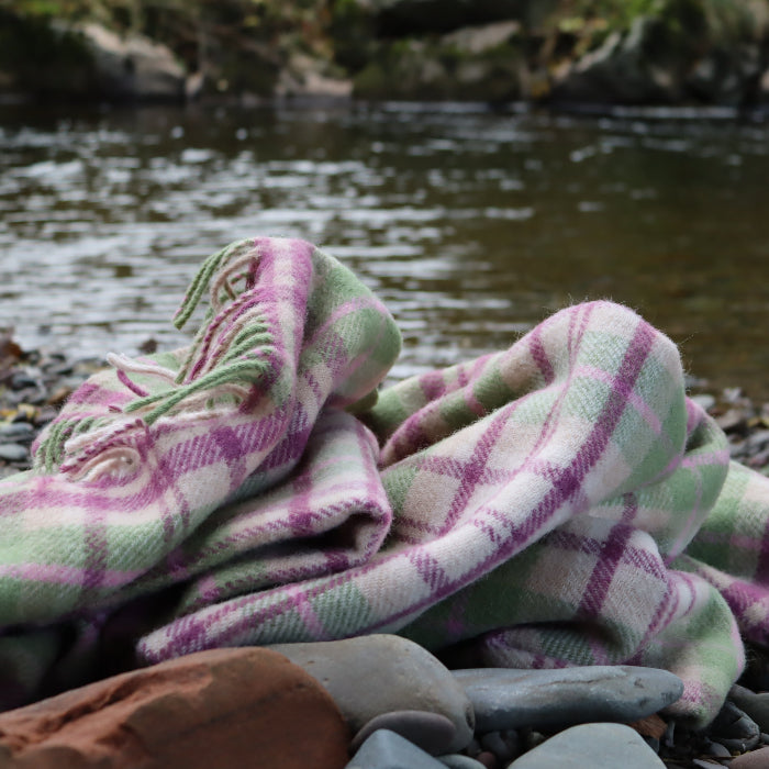 Blush pink, magenta, green and cream check wool blanket placed on a stone surface infront of a river 