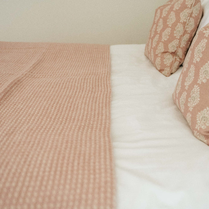 Rose coloured textured wool throw placed on top of white bedding with matching coloured cushions