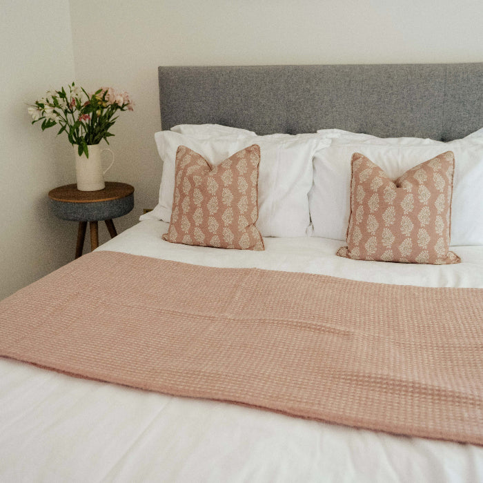 Grey bed with white bedding featuring a rose coloured textured wool throw and pillows in a bedroom setting with a white vase and flowers on a side table
