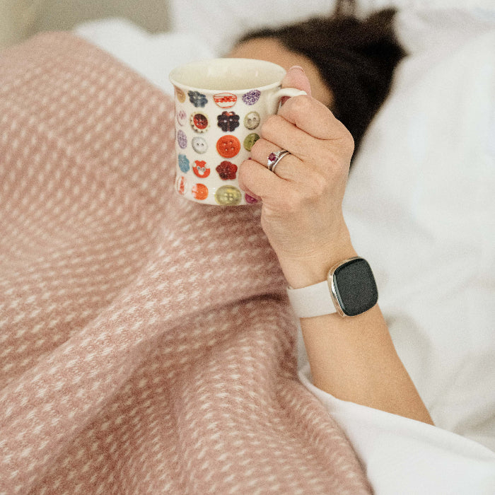 Person lying in bed holding a mug with a pattern, wearing a smartwatch, wrapped up in a rose coloured waffle textured wool throw. 