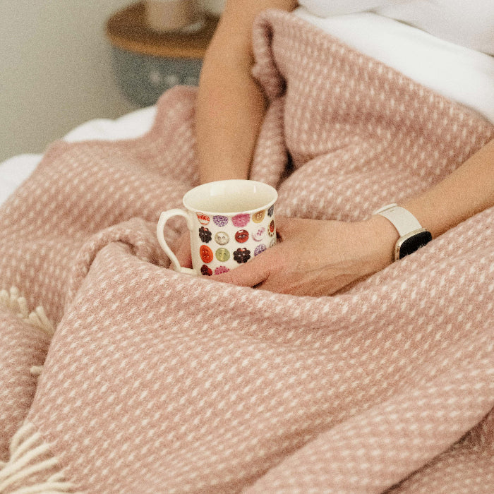 Person holding a mug under a rose coloured waffle textured blanket throw in a cosy bedroom setting
