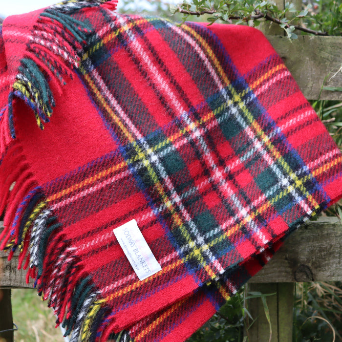 Red tartan wool blanket with colourful fringe and a Solway Blankets label, draped over a wooden fence ourdoors with a grassy background