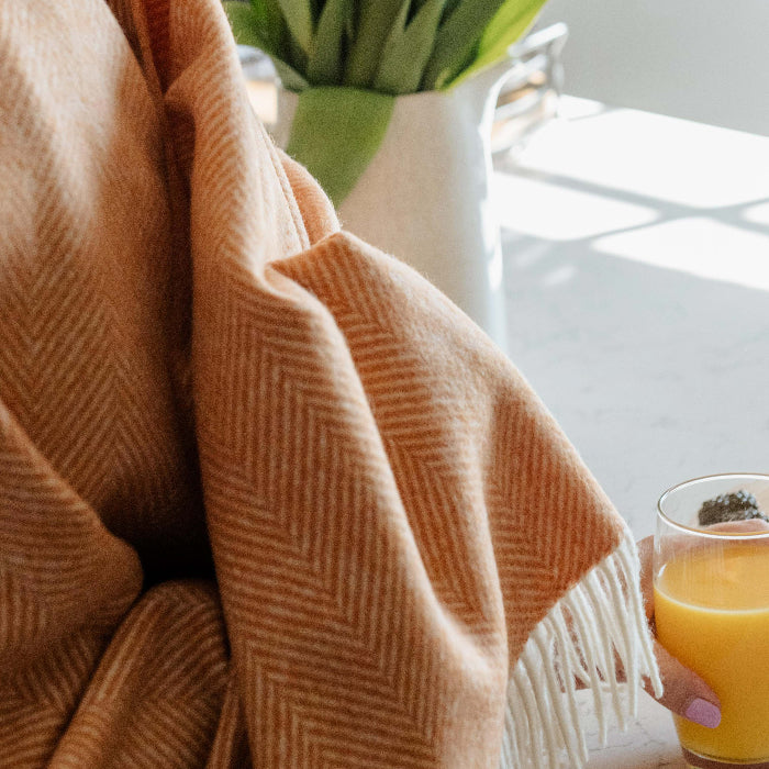 Person wrapped in a saffron orange herringbone blanket throw holding a glass of orange juice with flowers in the background