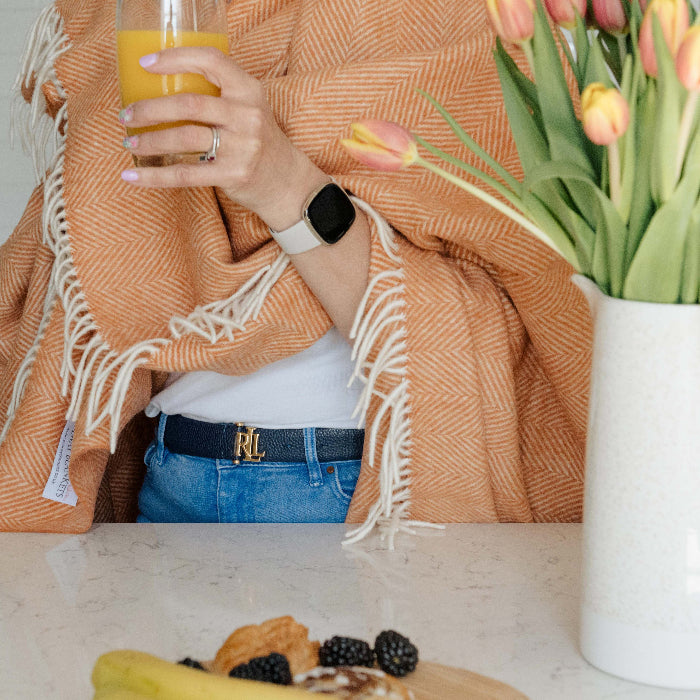 Person standing in a kitchen holding a glass of orange juice, with a saffron orange herringbone blanket throw wrapped around them. There is a vase with tulips on the table.