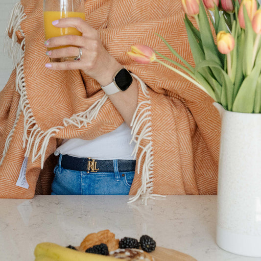 Person standing in a kitchen holding a glass of orange juice, with a saffron orange herringbone blanket throw wrapped around them. There is a vase with tulips on the table.