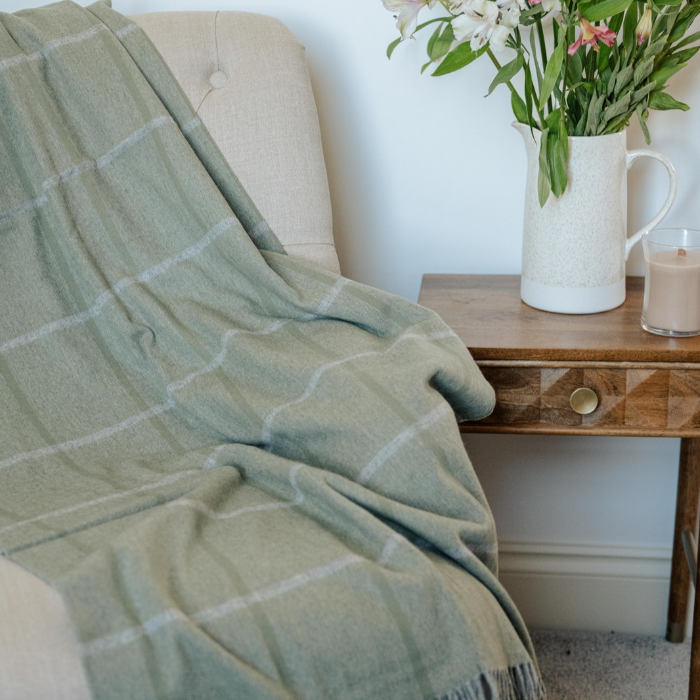 Eskdale Meadow pure cashmere throw in soft sage green with delicate windowpane check and twisted fringe draped over a cream tufted armchair next to a wooden side table holding a white pitcher of flowers and a lit candle.