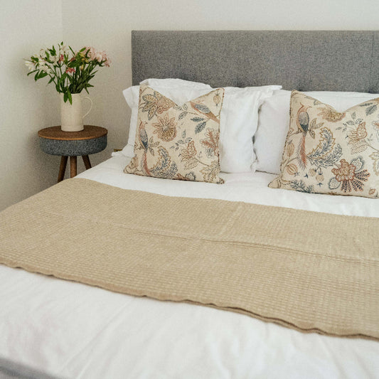 Bedroom with a bed featuring patterned pillows and a sandstone waffle textured wool throw, next to a small table with a vase of flowers.