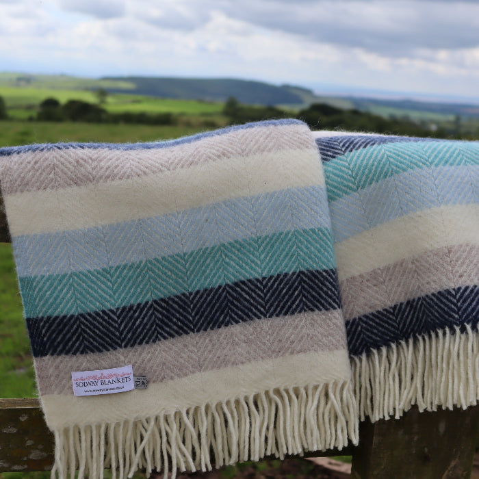 Cream, grey, navy, light blue and turquoise striped wool blanket with a 'Solway Blankets' label, folded over a wooden fence with a scenic background
