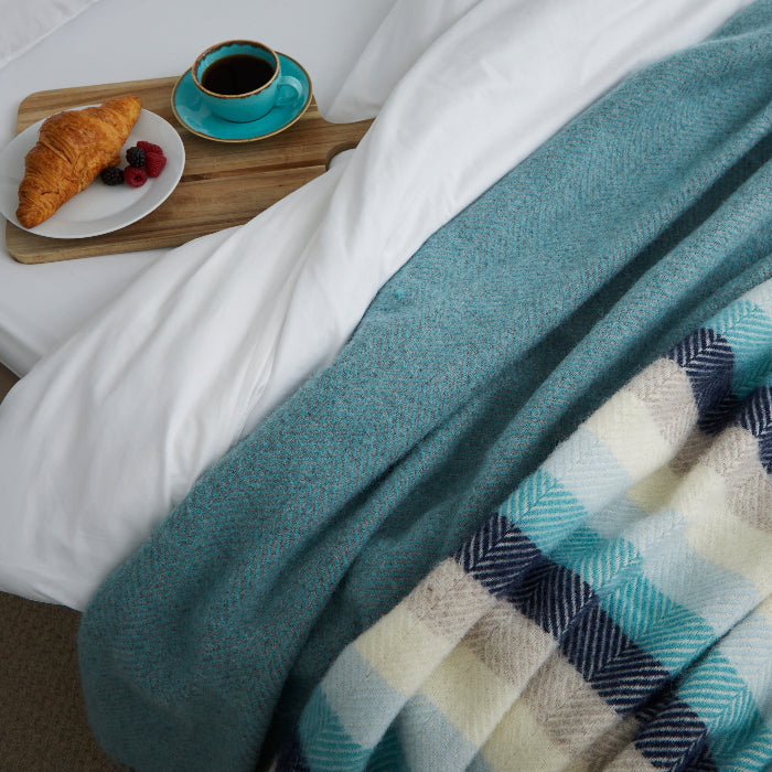 Cream, grey, navy, light blue and turquoise striped wool blanket draped over a bed with a wooden tray of coffee and pastries.