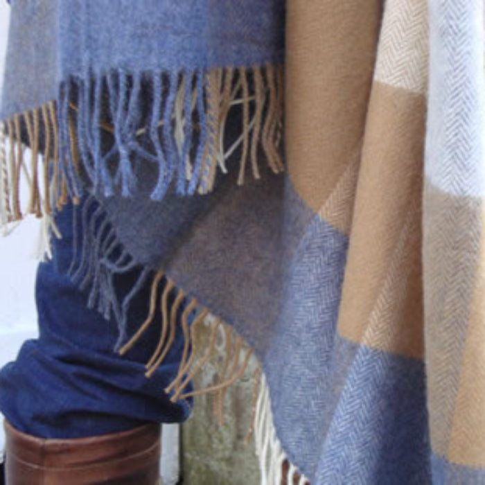 Plaid blue, tan and beige throw draped over a person's legs with fringes, against a stone wall background.