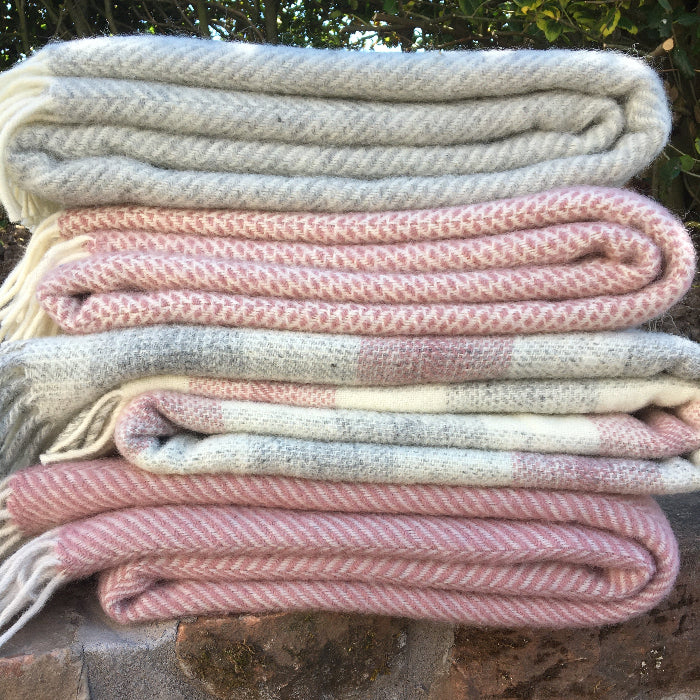 Set of four folded wool blankets in different patterns displaying pink and grey colours on a stone surface with greenery in the background