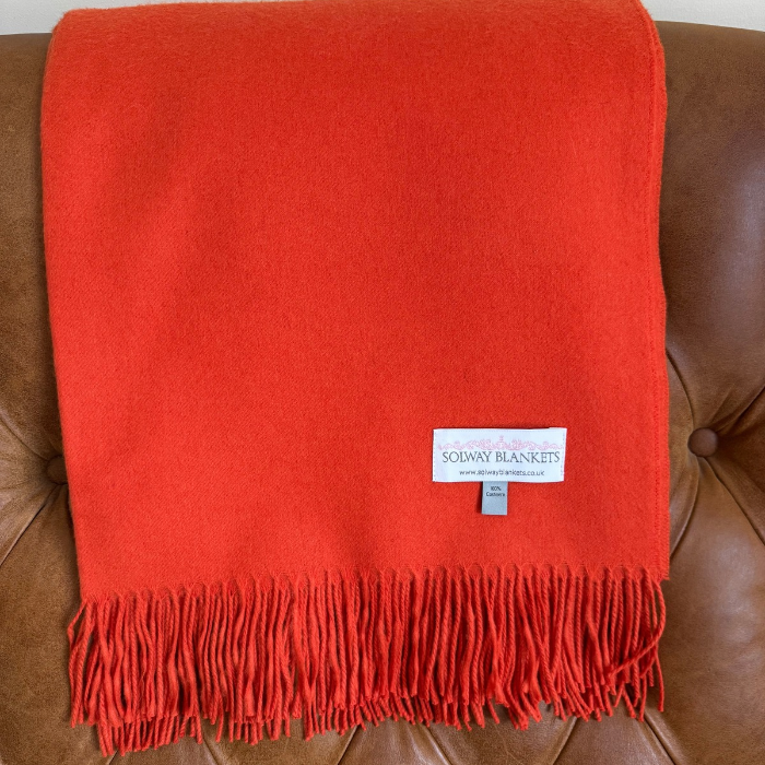 The vibrant orange Solway Sunset pure cashmere throw with matching fringe, woven in Scotland draped over a brown leather sofa