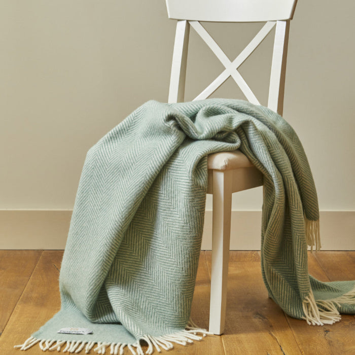 Spearmint green woolen blanket in a herringbone style, draped over a white chair against a beige wall.