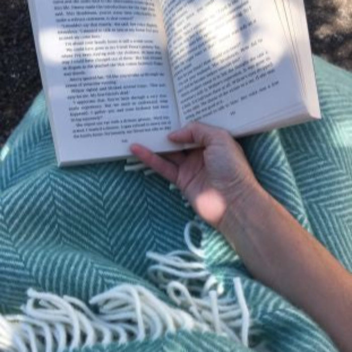 Person reading a book wrapped up in a green herringbone style woolen blanket, outdoors. 