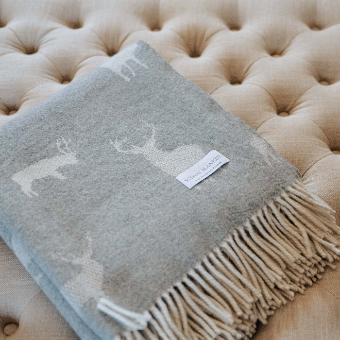 Grey blanket with deer pattern and fringe folded on a beige tufted ottoman