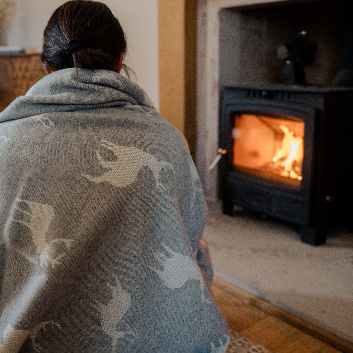 Person wrapped up in a cosy grey blanket with stags on sitting on the floor in front of a fireplace