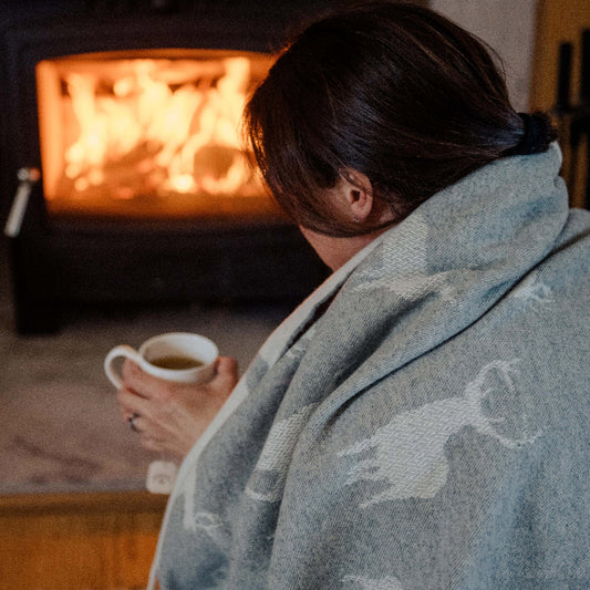 Person wrapped up in a stag covered blanket holding a mug in front of a fireplace