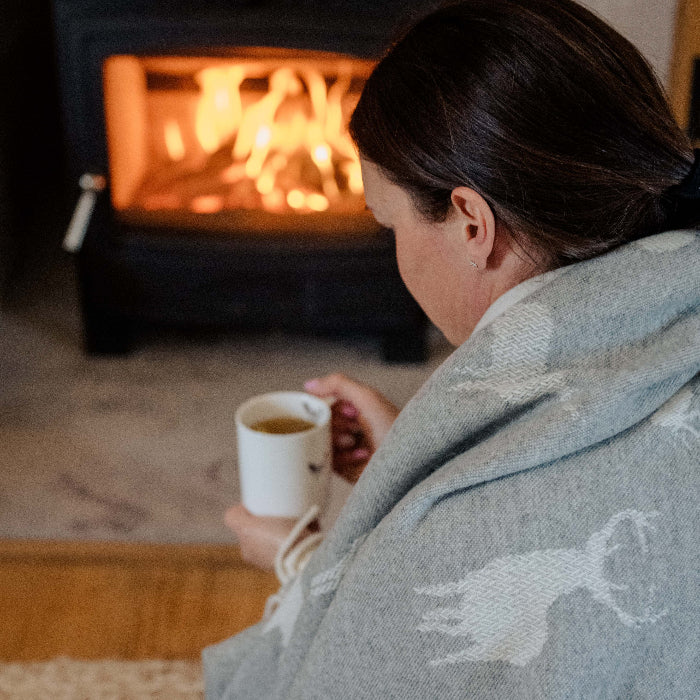 Close up of a person sitting by a fireplace holding a mug, wrapped in a cosy grey blanket with stag images on it