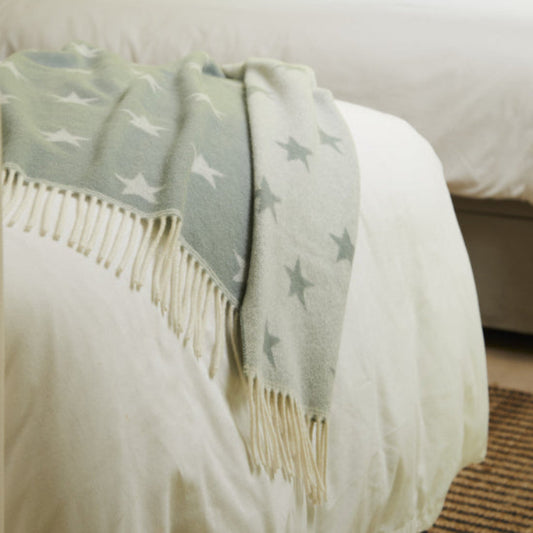 Bed with a Star Sea Green Merino Wool Baby Blanket, reversible featuring white star patterns and fringes draped over corner of the bed