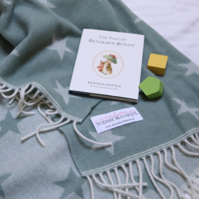 Star Sea Green Merino Wool Baby Blanket a muted green blanket with star patterns, a book, and colourful blocks on a white surface