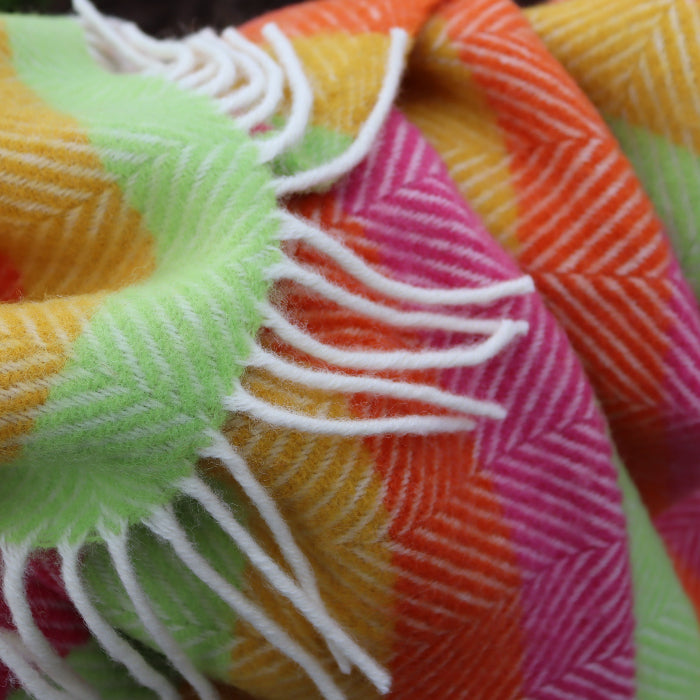 Close up of a colourful wool blanket with a herringbone pattern in four different colours - yellow, green, orange and pink with white fringes. 
