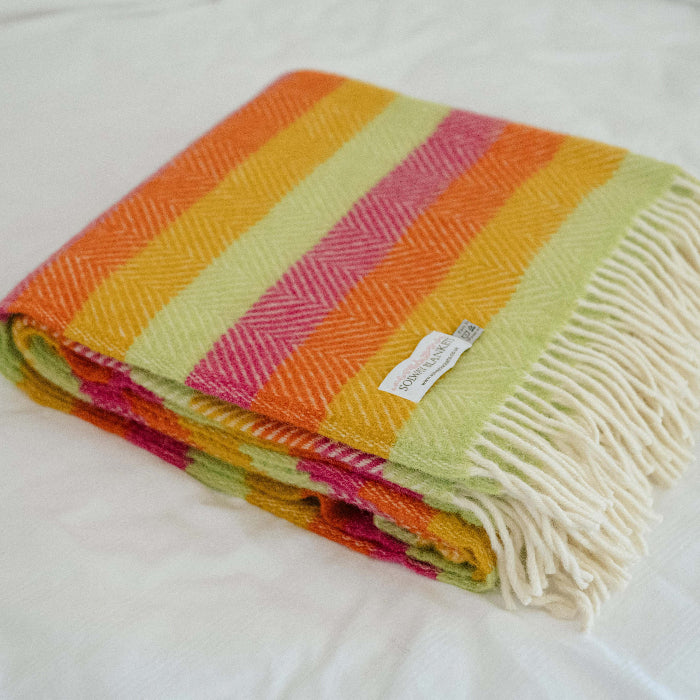 Colourful striped blanket with colours of  orange, yellow, pink and green with fringes and a visible 'Solway Blankets' label, folded neatly on a white bed