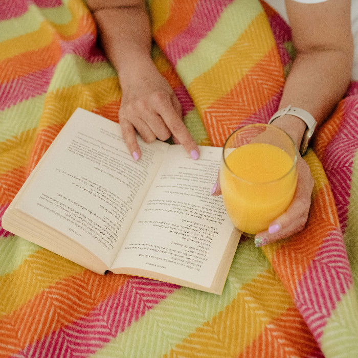 Person lying on a white bed, reading a book and holding a glass of orange juice leaning on a colourful striped wool blanket