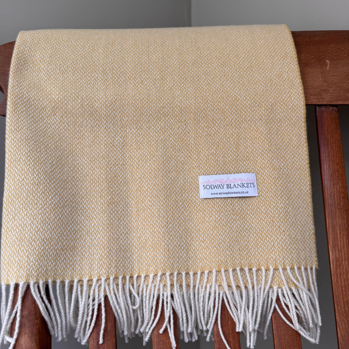 A folded beautifully woven cashmere baby blanket in a warm cream-yellow colour with white fringe, draped over a wooden chair. A white brand label reading 'SOLWAY BLANKETS' is sewn onto the front. Made in Britain