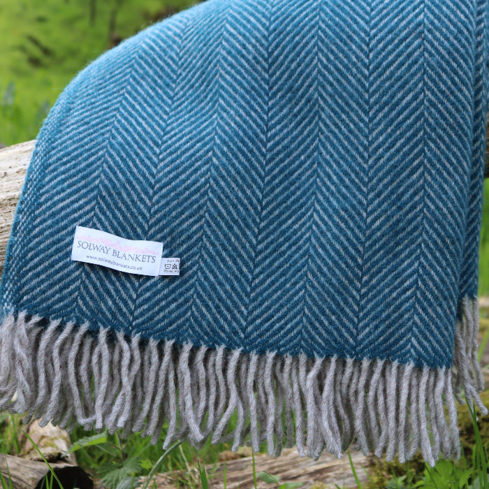 Teal blue herringbone blanket with fringes featuring a 'Solway Blankets' label, draped over a log in a natural setting