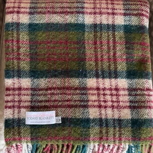 A close-up of the woven Thistleberry Tartan plaid blanket with fringe at the bottom. The pattern mixes cream, olive green, dark teal-blue, and magenta-pink in a tartan design.