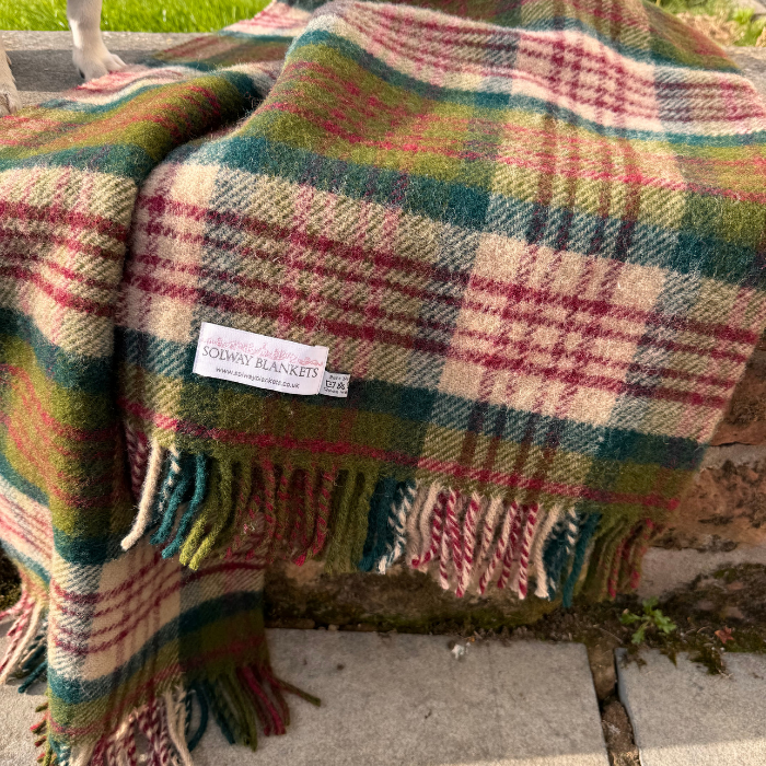 A close-up of a soft woven Thistleberry Tartan knee rug with fringe, shown draped over a rustic brick surface. The blanket features check design of rich red, dark green, teal and beige.