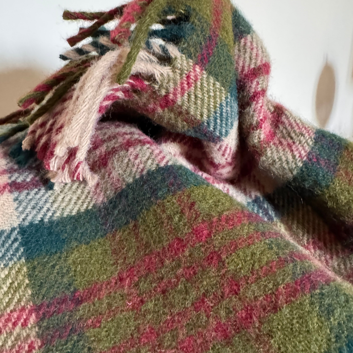 A close-up of a plaid wool blanket with fringe, featuring Thistleberry check design with moss green, teal, berry red and beige.