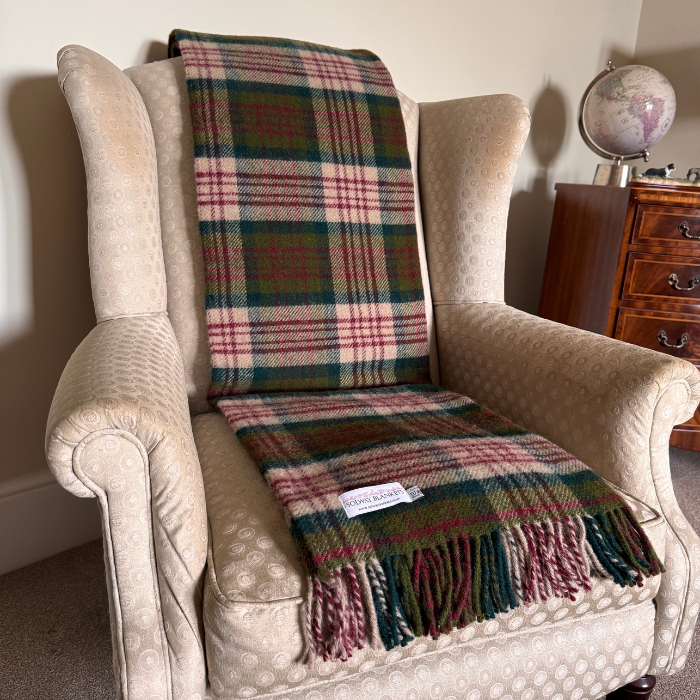 A close-up of the Thistleberry Tartan Check Pure Wool Blanket with fringe, featuring red, dark green, beige tones, draped over a neutral arm chair in a cosy living room