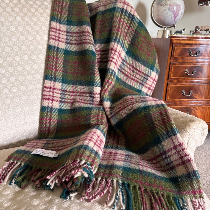 A close-up of a woven plaid Thistleberry tartan wool blanket draped over an arm chair, showing a multicolour check plaid pattern with red, beige and green, finished with fringed edges and a visible Solway Blankets label.