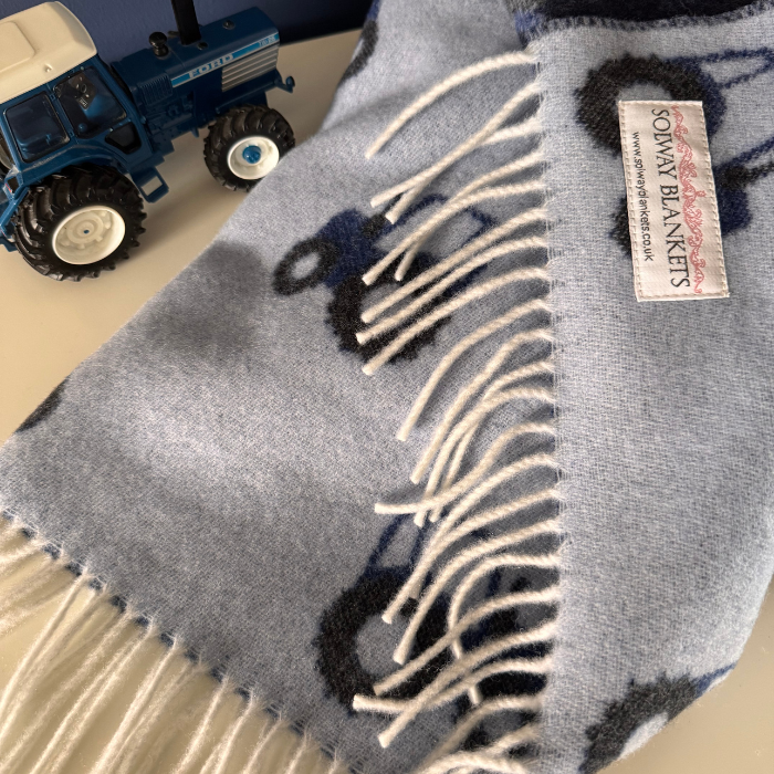 A close-up of a light blue woven merino baby blanket with white fringe, featuring a dark blue and black tractor pattern. A white label reading 'SOLWAY BLANKETS' is visible, and a blue toy tractor sits in the top-left corner on a light surface.