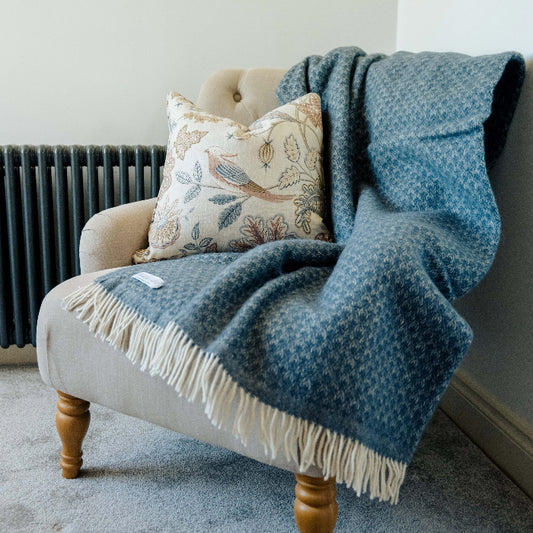 Twilight blue wool throw with fringe, draped over a beige chair with wooden legs and patterned cushion placed on top. 